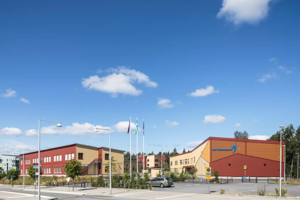Bild tagen från gatan på skolan som har en fasad i röda och gula partier. I förgrunden syns skolans parkering där det står en bil.