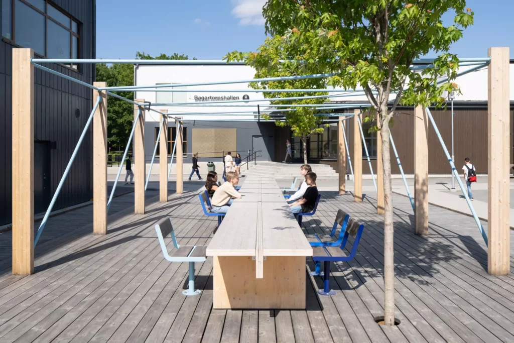 Fyra elever sitter vid ett avlångt bord utomhus under en pergola. Några gröna träd syns till höger i bild.