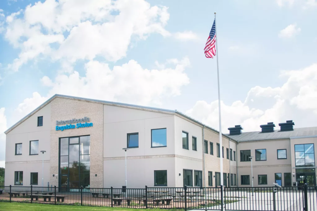 En vit tvåvåningsbyggnad byggd i vinkel. På kortsidans gavel står det Engelska Internationella Skolan. En amerikans flagga vajar i en flaggstång på gården.