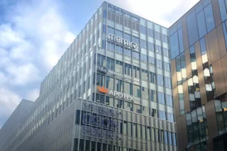 En modern och nybyggd fastighet på åtta våningar med stora fönsterpartier syns på fotot. Flers skyltar med företagslogotyper som apoteket och Memira syns på fasaden.