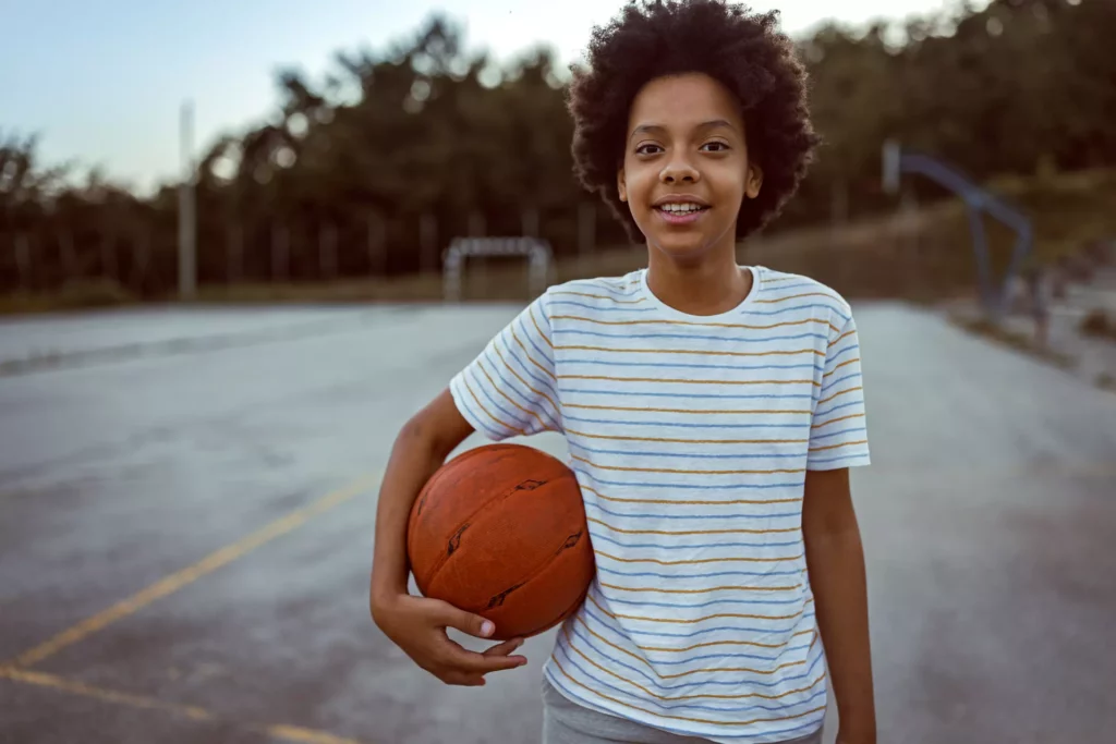 Ett barn i randig t-shirt står på en spelplan med en basketboll under ena armen