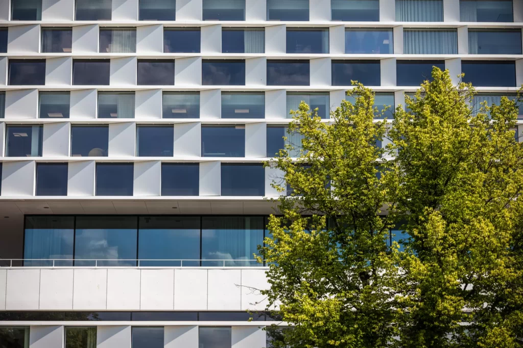 Mälardalens universitet ha en fasad med många fönster och vita partier däremellan. I förgrunden står ett stort grönt träd. Längst ner i bild ses en våning i glas med balkong utanför.