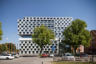 Mälardalens universitet i Eskilstuna har en fasad med många fönster och vita partier däremellan. I förgrunden en parkering med flera bilar och några stora, gröna träd.