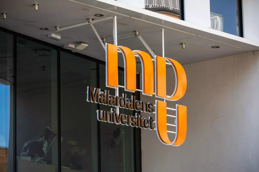 En skylt i entrén med Mälardalens universitets logga i orange. Skylten hänger i luften och är fäst i taket.