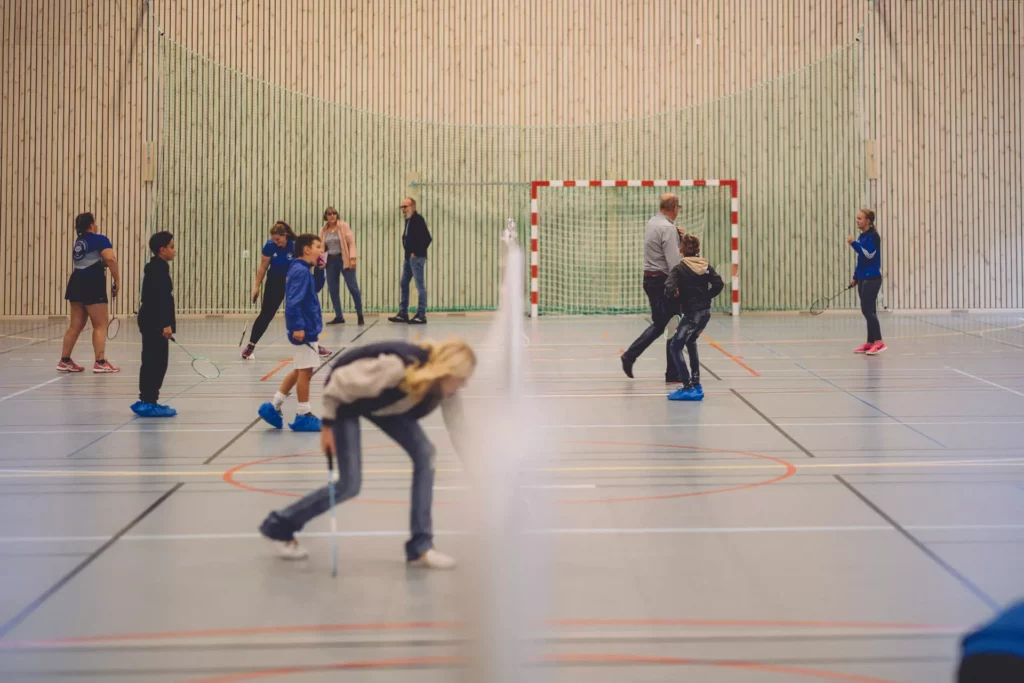 Elever syns spela badminton inne i idrottshallen. Väggen är klädd med ljusa träribbor och golvet har märkning för olika sporter.