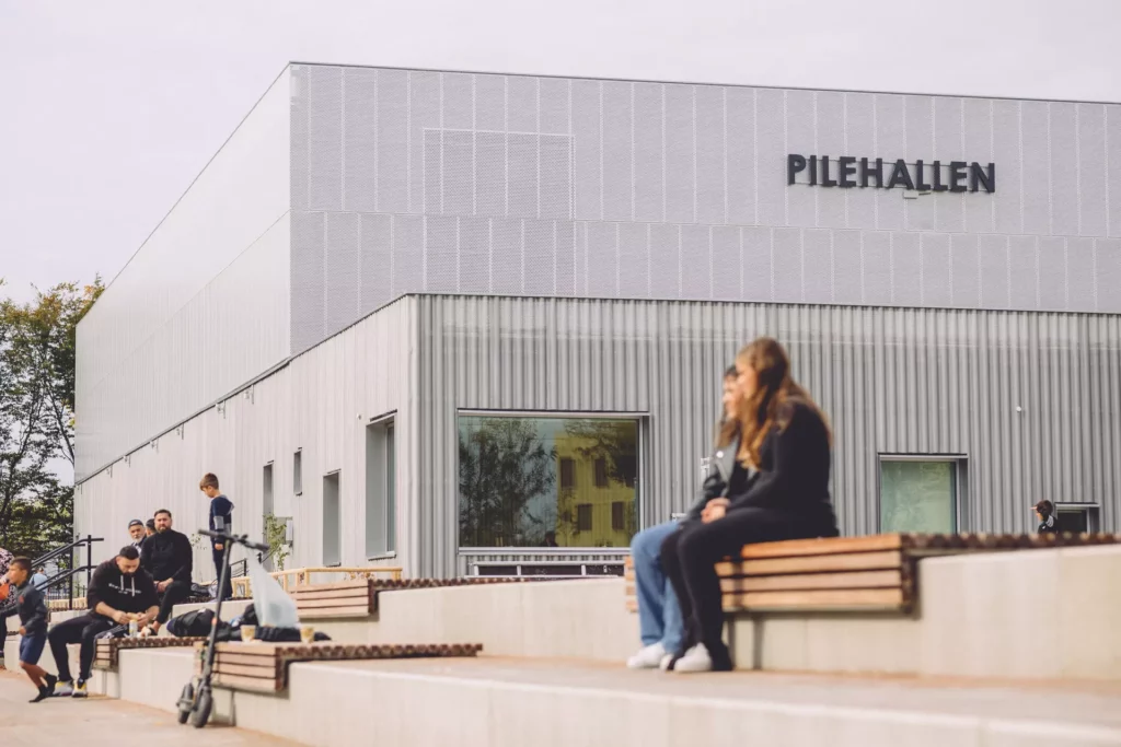 Bild på Pilehallen tagen utifrån trapporna med sittplatser som finns utanför hallen. Flera människor sitter på sittplatserna av träribbor.