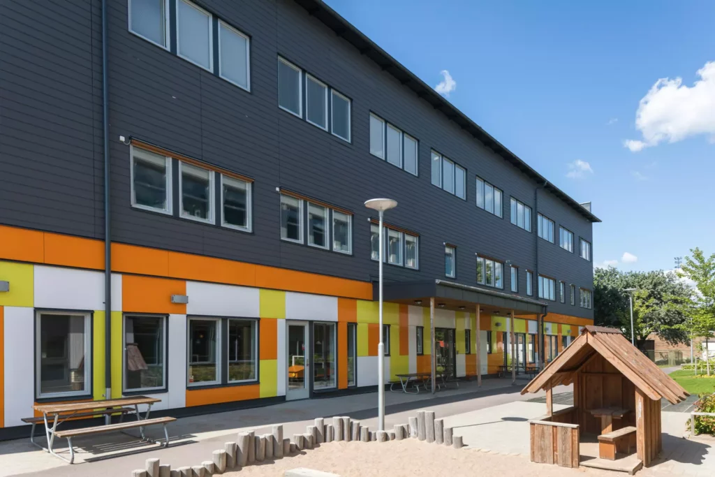 Lekfulla färger i vitt, gult och orange pryder den nedersta våningen av fastigheten Rosenhill 12. I förgrunden syns en sandlåda och en lekstuga. De över två våningarna har fasad i mörkgrått.