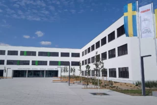 Skolan i Zossen är vit och byggd i tre våningar. På fotot syns entrén till skolan. Den svenska flaggan, den tyska och en Hemsöflagga syns till höger i bild.