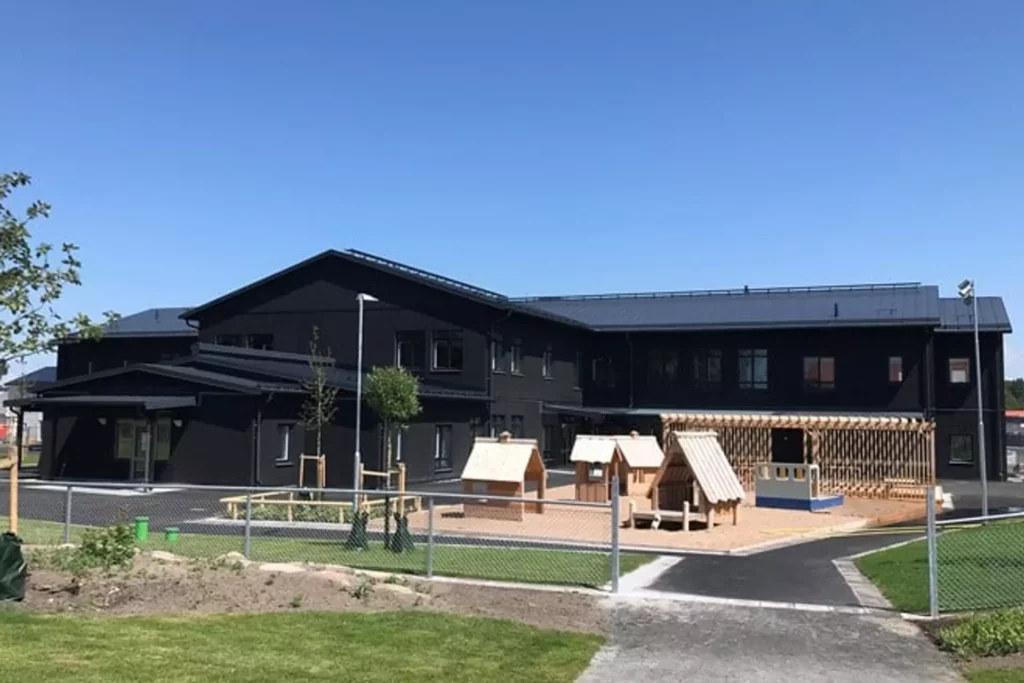 Förskola fotad utifrån. En mörk träbyggnad med en fin lekpark i ljust trä framför.