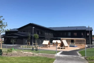 Förskola fotad utifrån. En mörk träbyggnad med en fin lekpark i ljust trä framför.