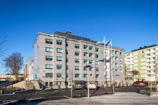 Ett sex våningar högt hus med grå, robust fasad är polishuset mi Rinkeby. I förgrunden några flafggor med polisens emblem och en parkering.