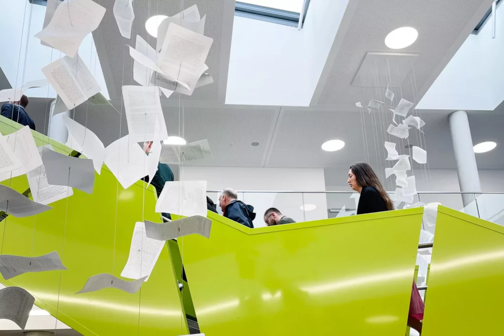 En grön trappa i Hemsös skola i Zossen, Tyskland. Från taket hänger ett konstverk i form av pappersark i snören med text på i olika nivåer.