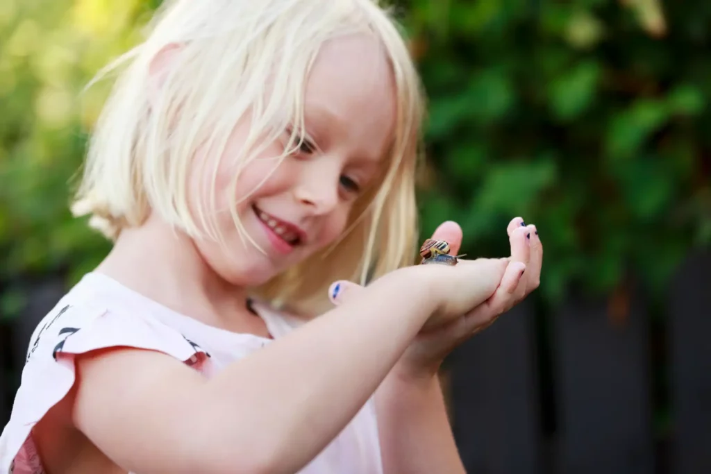 En blond flicka håller upp sin hand där det sitter en liten snigel, hon tittar på snigeln och ler.