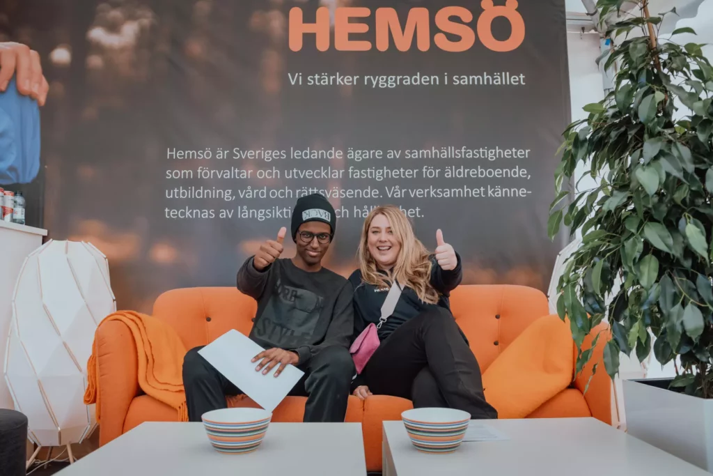 A young man and woman are sitting in an orange sofa in front of a Hemsö roll up ang doing their thumbs up.